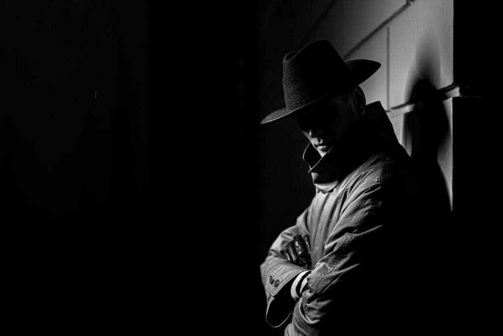 A black and white photograph capturing a mysterious figure draped in shadows, wearing a hat and a trench coat