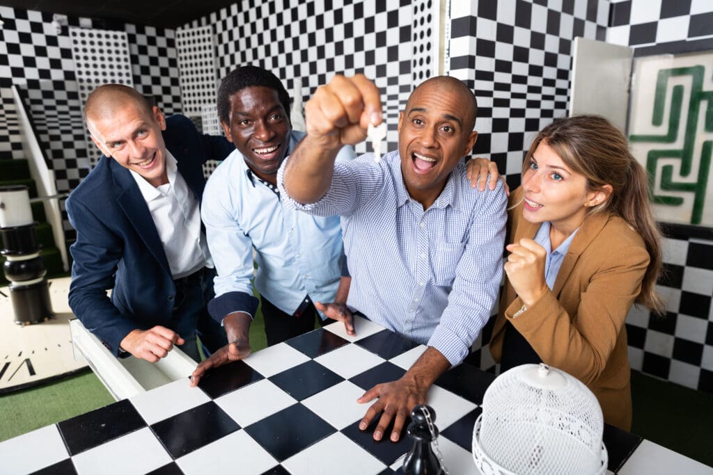 A diverse group of four colleagues working together in an escape room over a giant chess-themed game table, expressing teamwork in a playful setting.