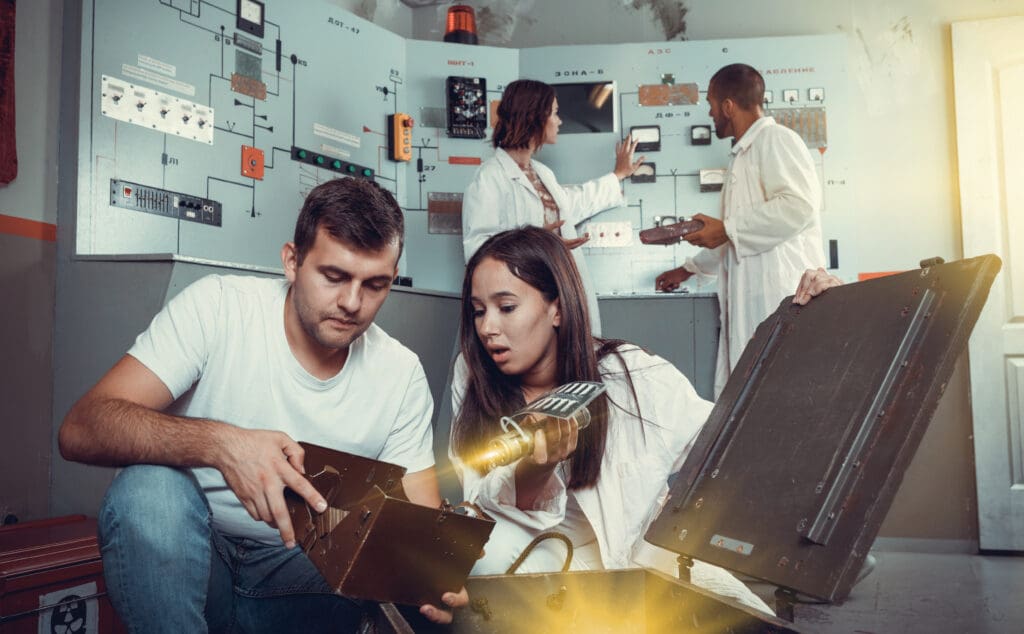 Two young adults, a man and a woman, closely examining and working with a piece of technical equipment in an escape room, with other team members actively engaged in the background.