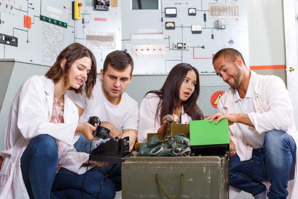 A diverse group of four young adults solving an escape room puzzle, examining and discussing together in a control room filled with technical diagrams and electrical panels.