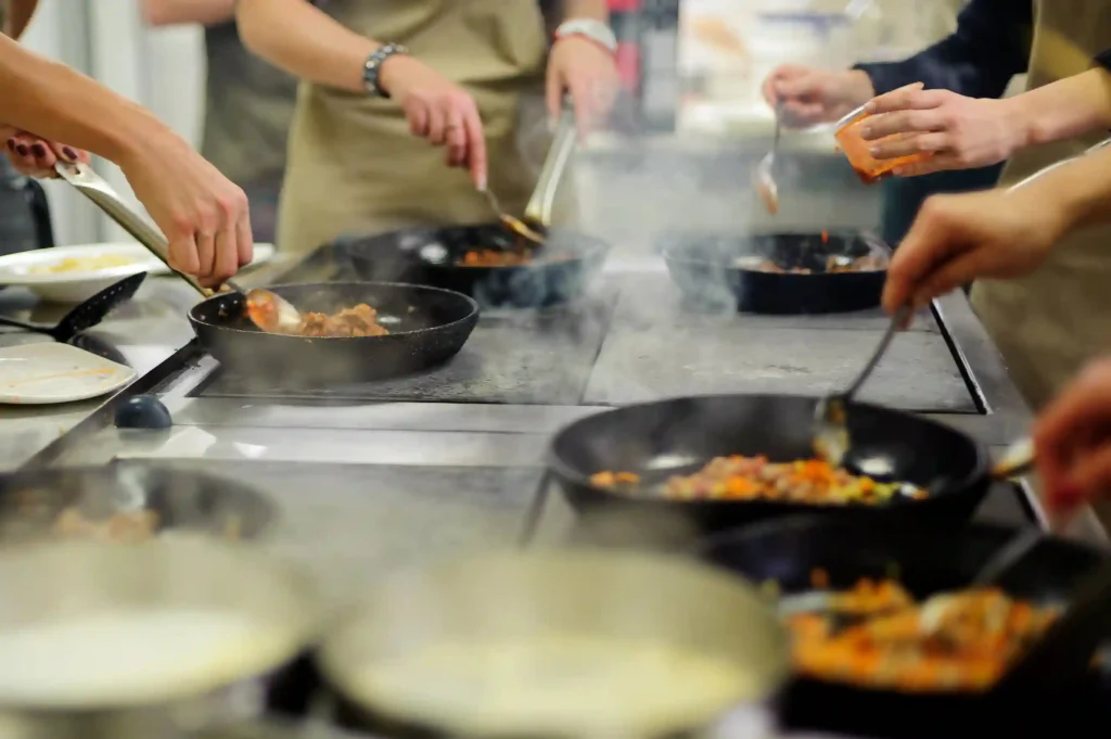 People participating in a group cooking activity, stirring food in pans on a stove during a cooking competition.