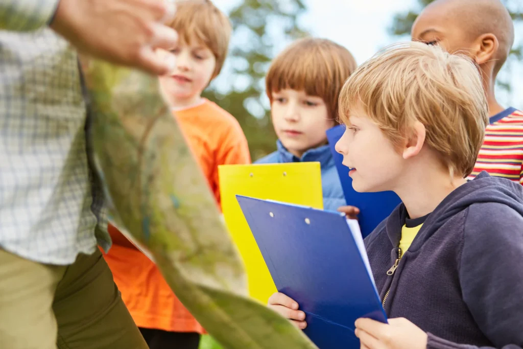 Group of children outdoors participating in a scavenger hunt, holding colourful clipboards and examining a map.