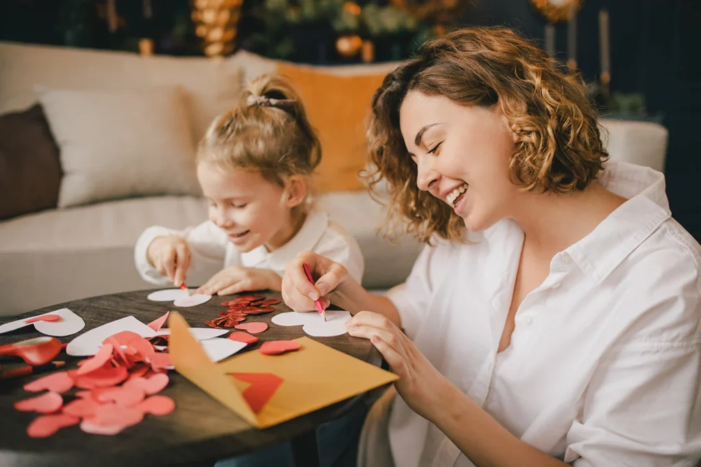 Mother and daughter sitting at a table crafting paper hearts together, sharing smiles and laughter.