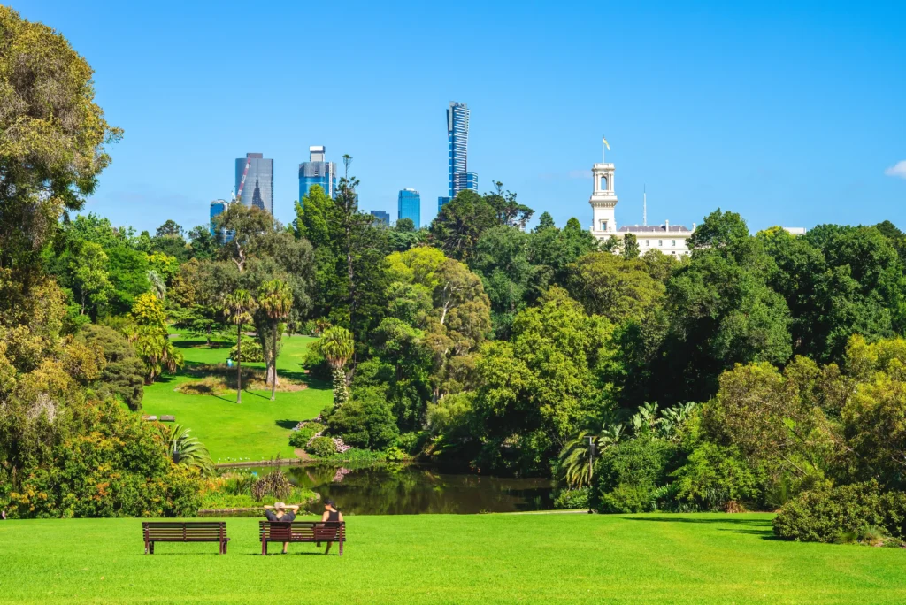 A scenic view of Melbourne's Royal Botanic Gardens with lush greenery, benches, a pond, and city buildings in the background."