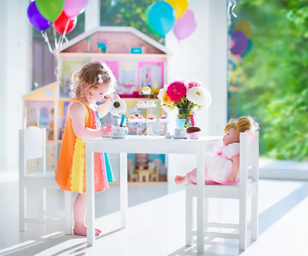 Young child hosting a tea party with a doll, surrounded by colourful balloons, flowers, and a dollhouse in a bright and cheerful room.