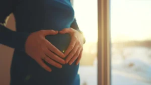 Close-up of a pregnant woman holding her belly, standing by a window with sunlight streaming in, symbolising comfort and care.